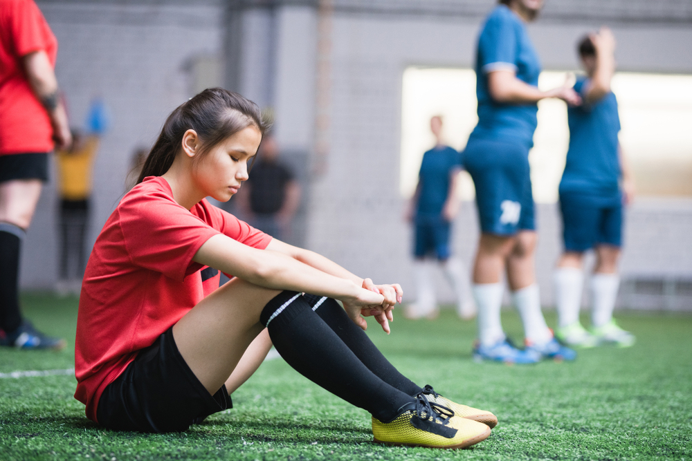 Tired young athlete sitting on the field after sports activity, illustrating the need for rest and recovery in preventing sports injuries