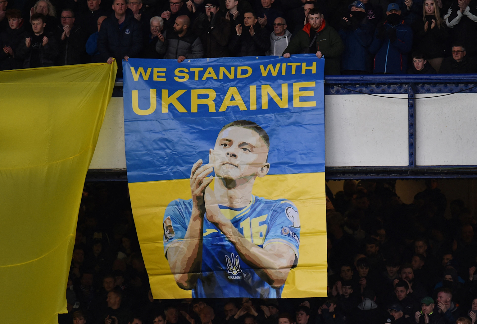 Supporters in a stadium display a banner with the message 'We stand with Ukraine' featuring the Ukrainian national football team colors and a player illustration