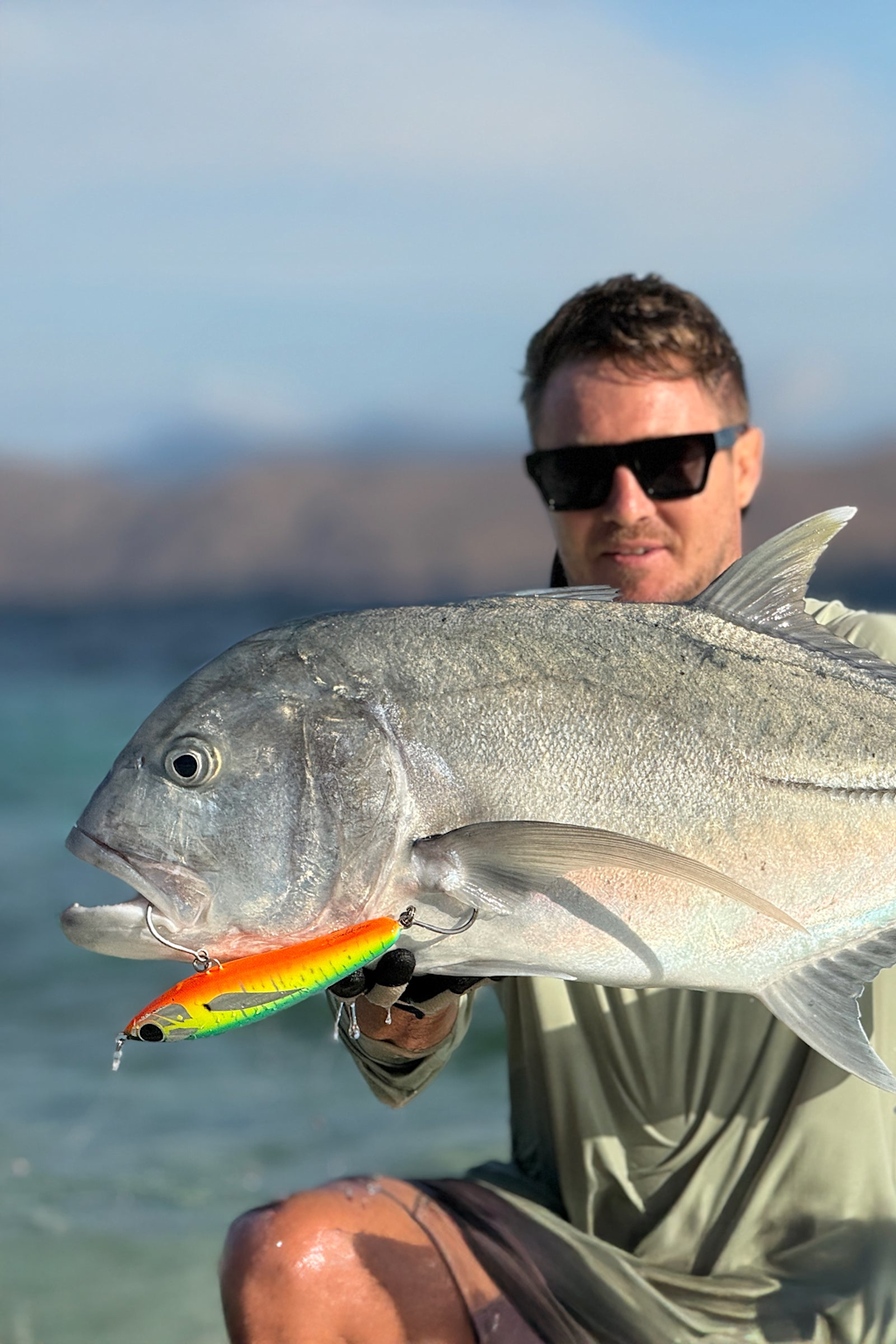 A person holding a large fish caught using a colorful fishing lure near a scenic water body