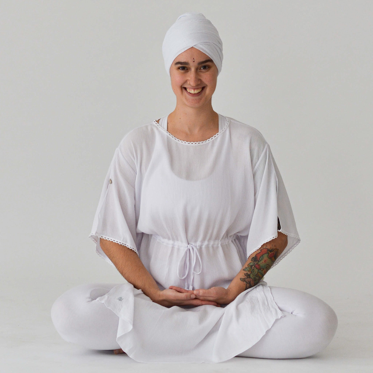 A woman in traditional white Kundalini yoga clothing sits in meditation with a calm smile in a minimalist setting