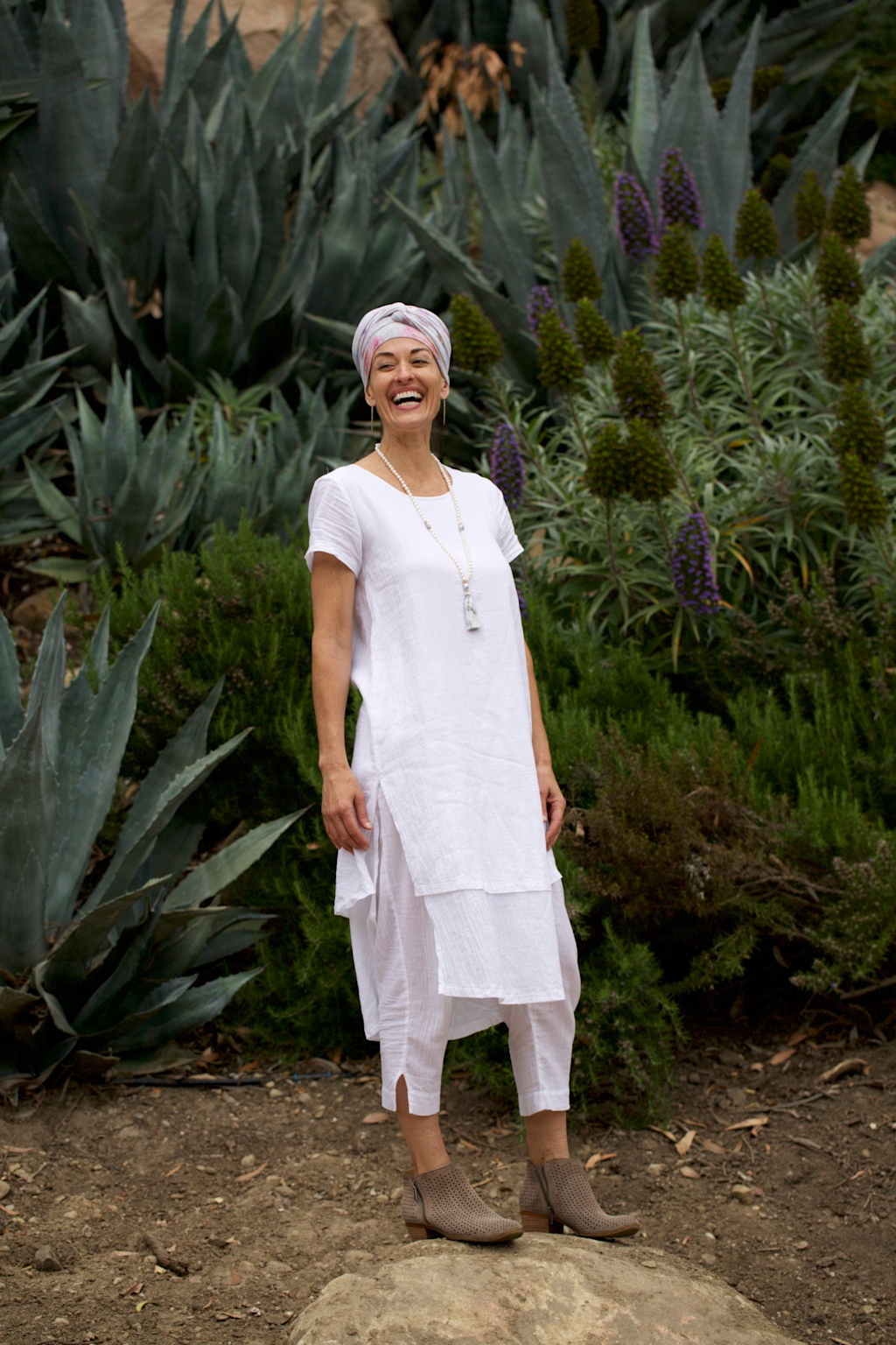 A woman in white Kundalini yoga attire smiling outdoors among green plants and rocks