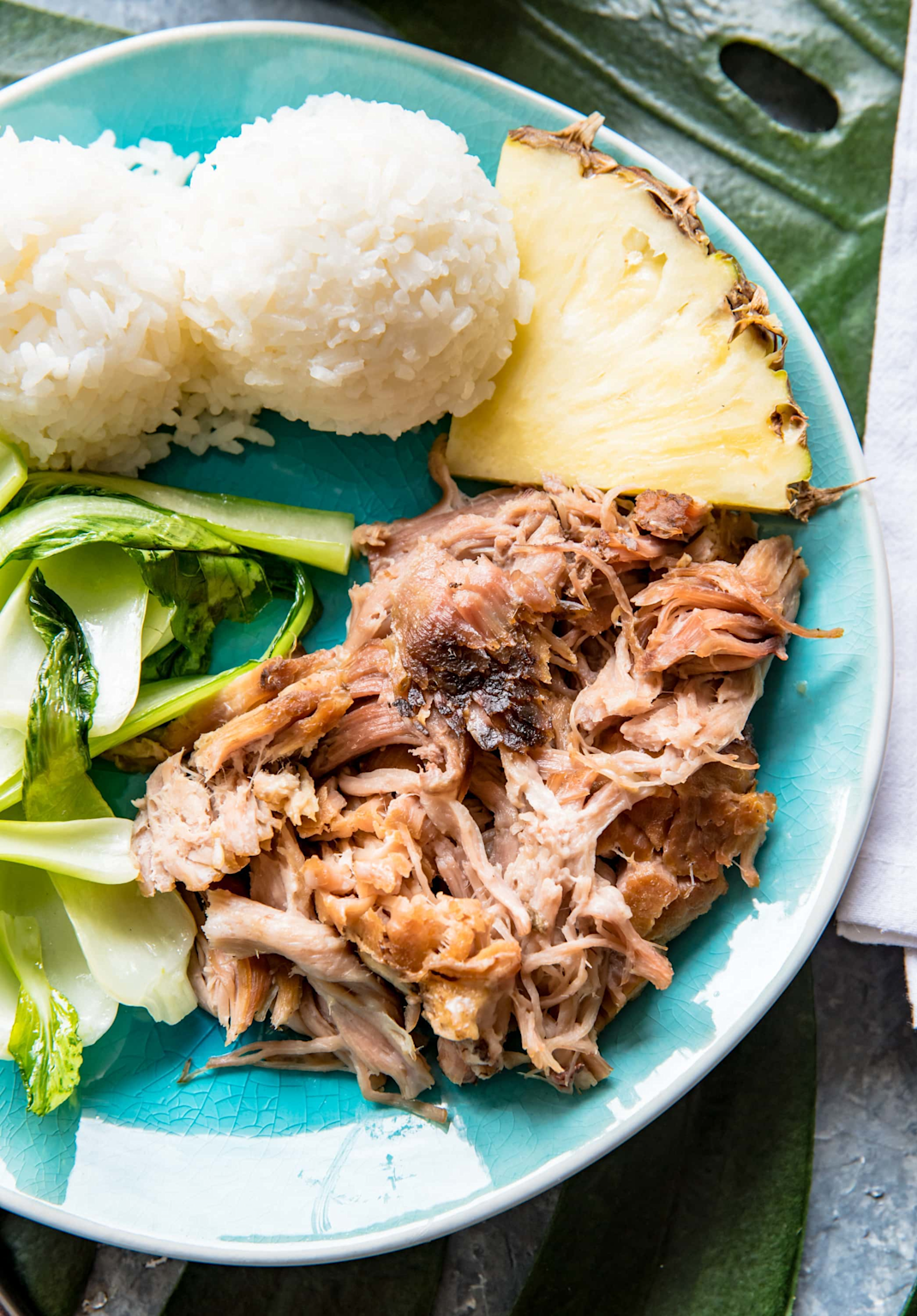 Classic Hawaiian plate lunch with tender Kalua pork, steamed rice, fresh pineapple, and leafy greens on a blue plate