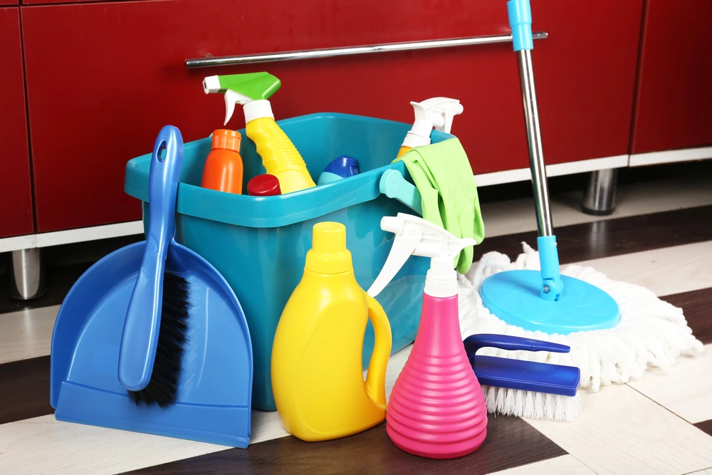 A set of colorful cleaning tools and supplies including spray bottles, a dustpan and brush, mop, and cloths arranged neatly