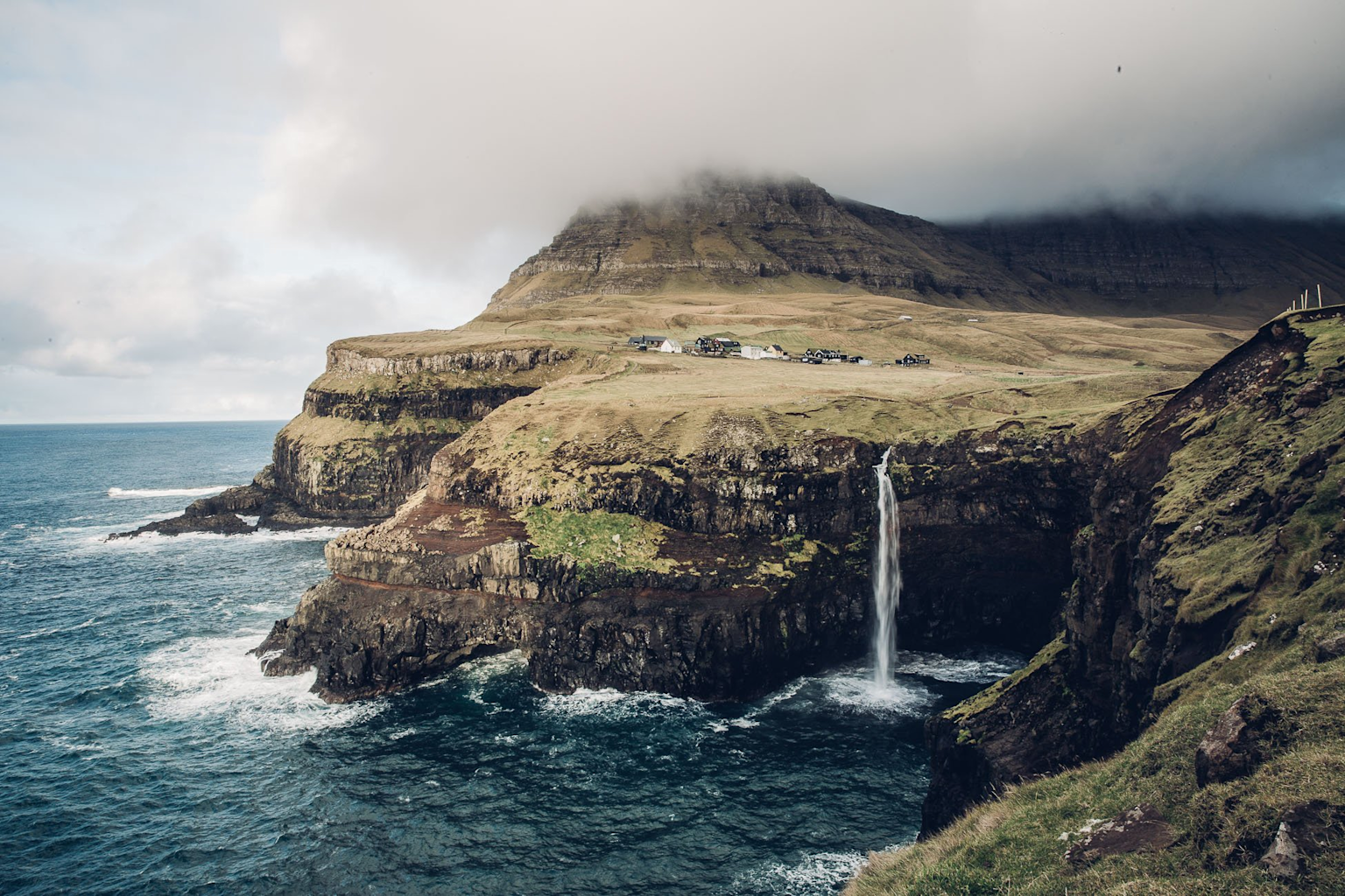 Mulafossur waterfall cascading into the ocean on the Faroe Islands with dramatic cliffs and low clouds