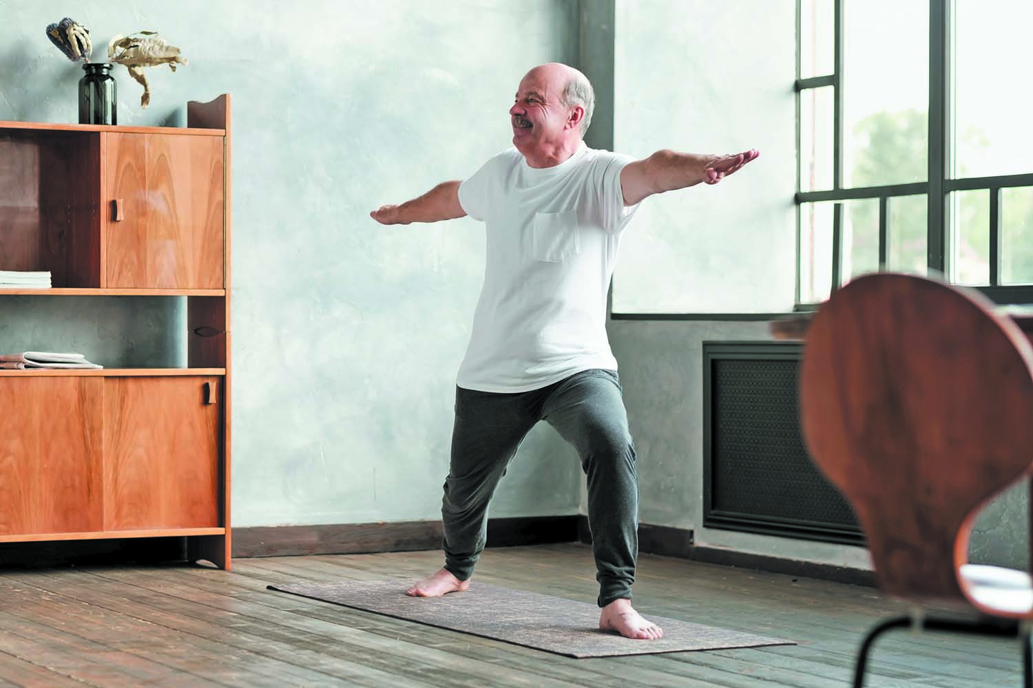 An elderly man practicing warrior II yoga pose indoors as part of cardiac rehabilitation therapy