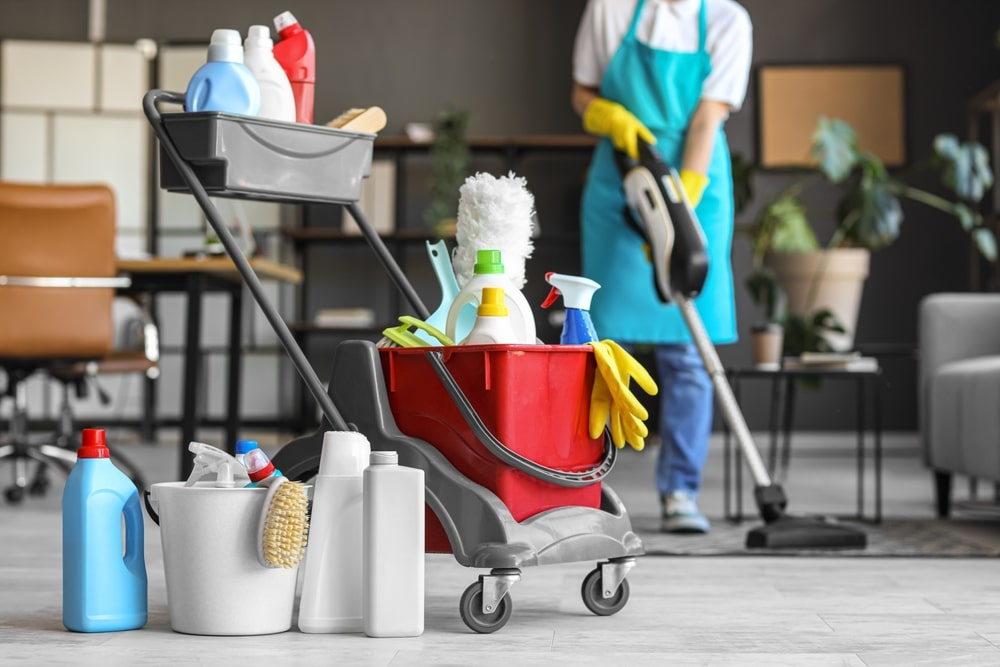 Professional commercial cleaning supplies and equipment including mop bucket, cleaning liquids, brushes, gloves, and vacuum cleaner in use