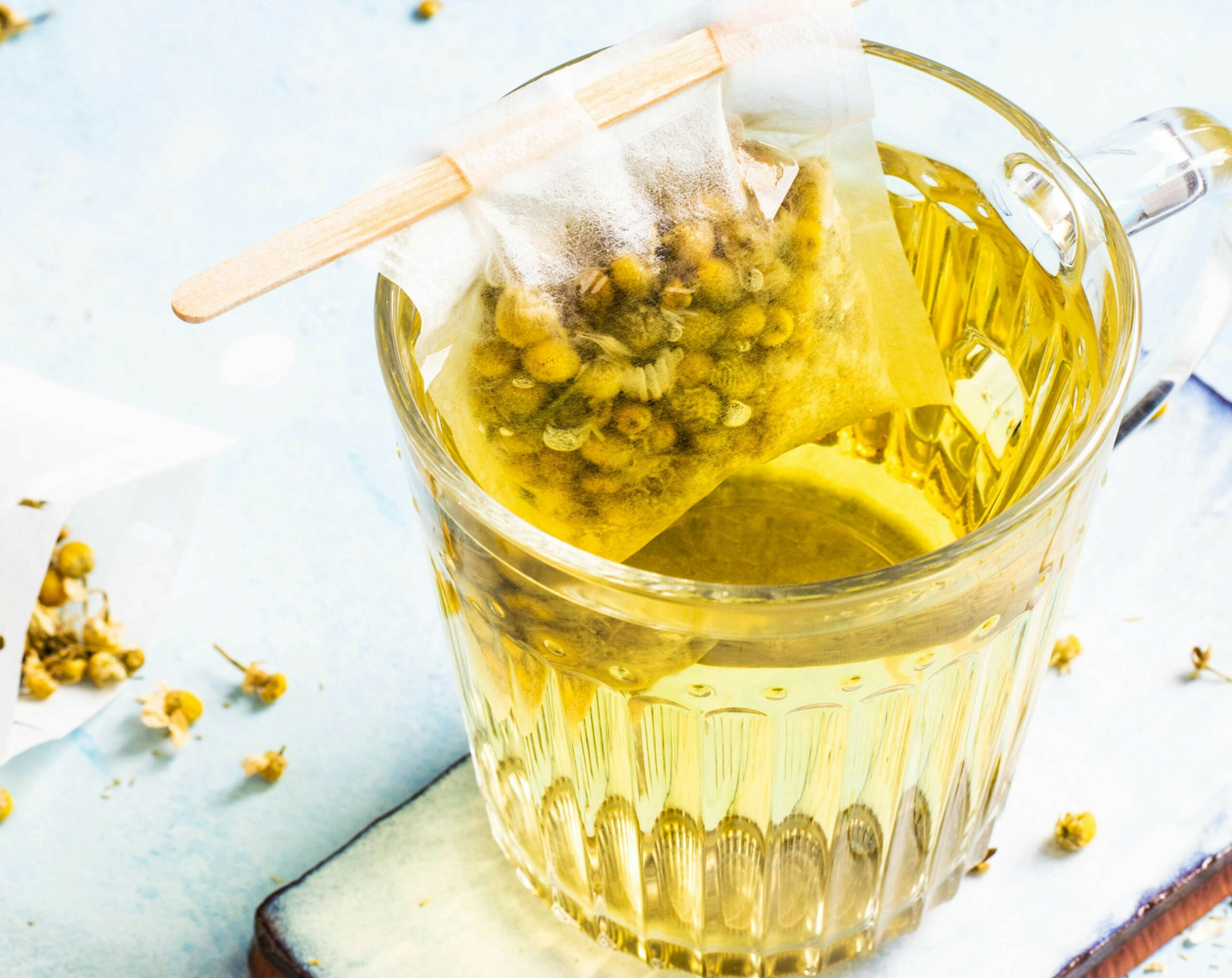 A clear glass cup of chamomile tea with a tea bag steeping, surrounded by chamomile flowers on a light surface