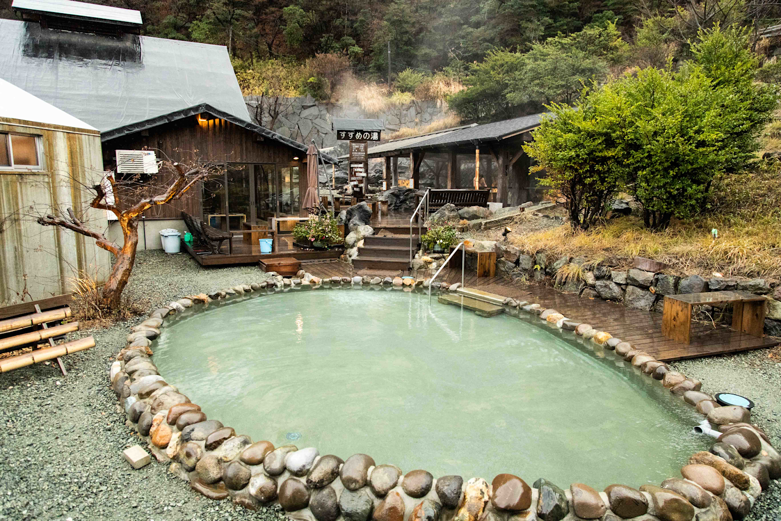 A tranquil outdoor hot spring (onsen) in Kyushu, Japan, surrounded by natural volcanic landscapes and traditional wooden structures