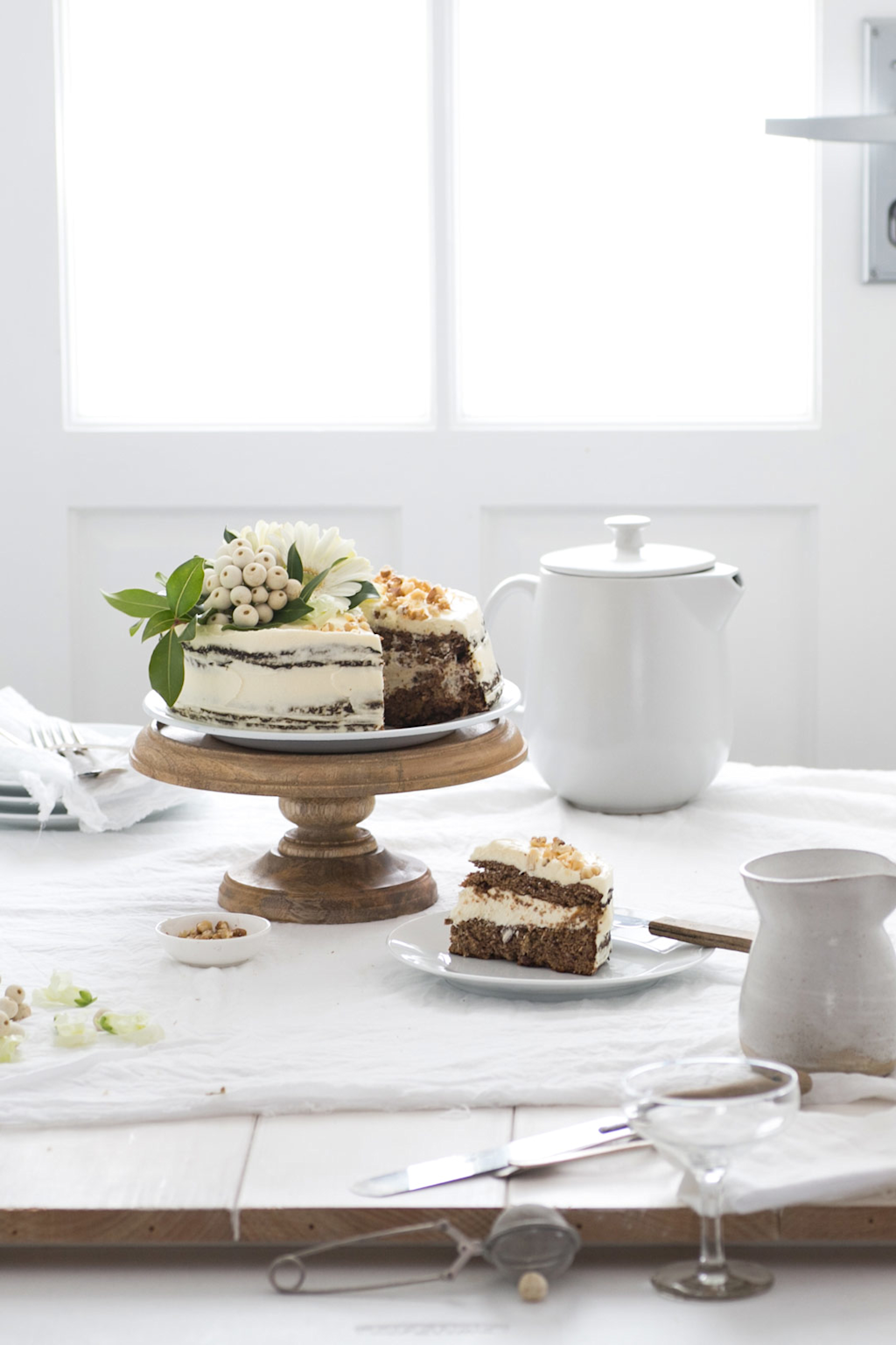 A cake on a wooden stand by a window with natural light, surrounded by simple props for an inviting food photography setup.