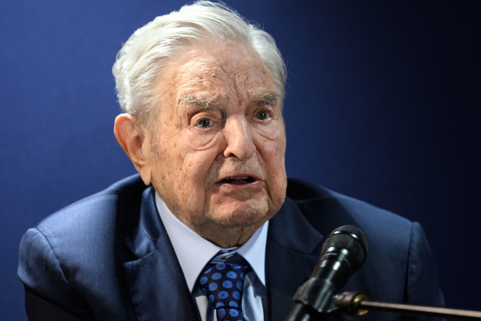 Elderly man in a formal suit speaking into a microphone, likely George Soros, associated with progressive funding initiatives