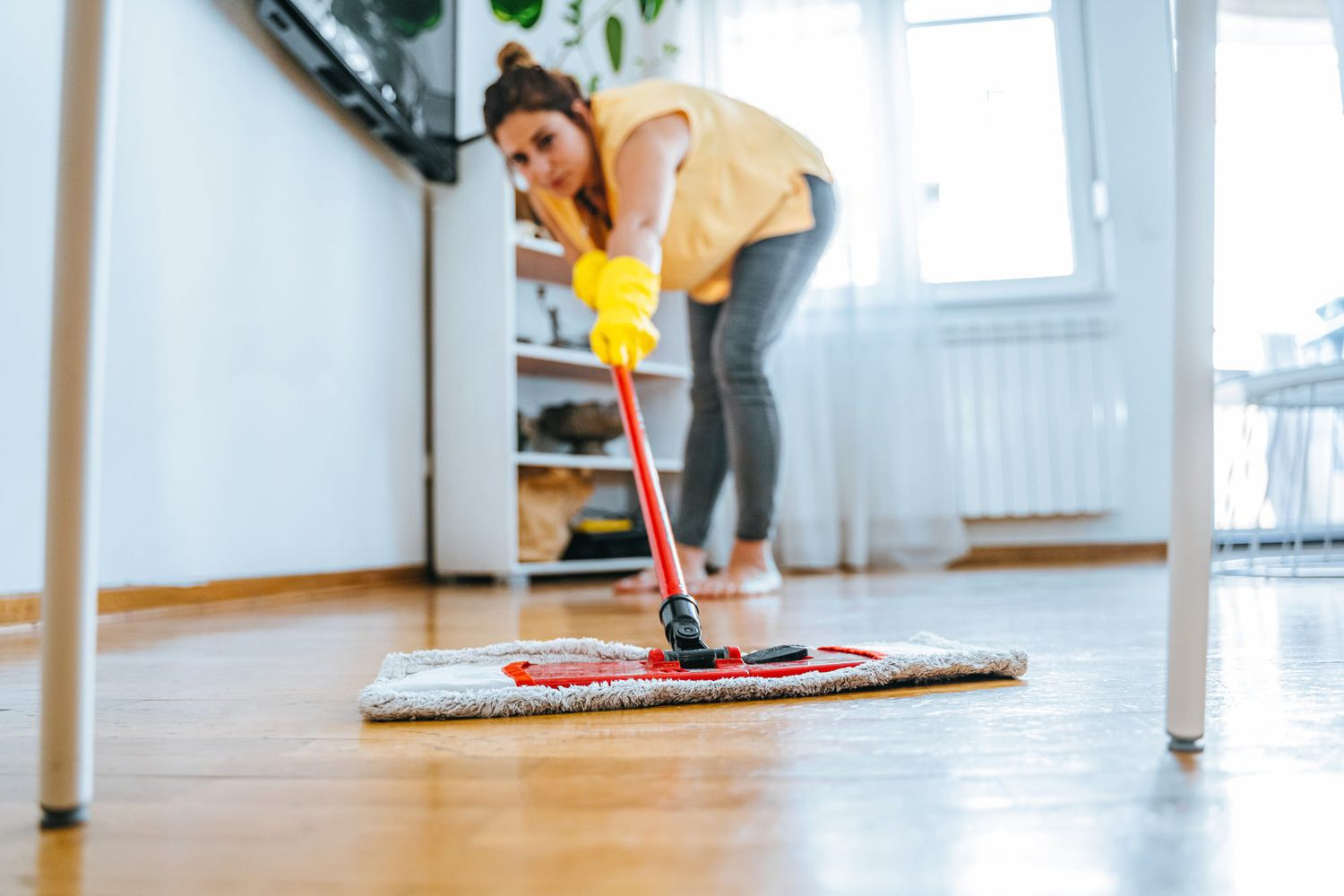 A person cleaning a wooden floor with a mop and microfiber cloth, wearing yellow gloves