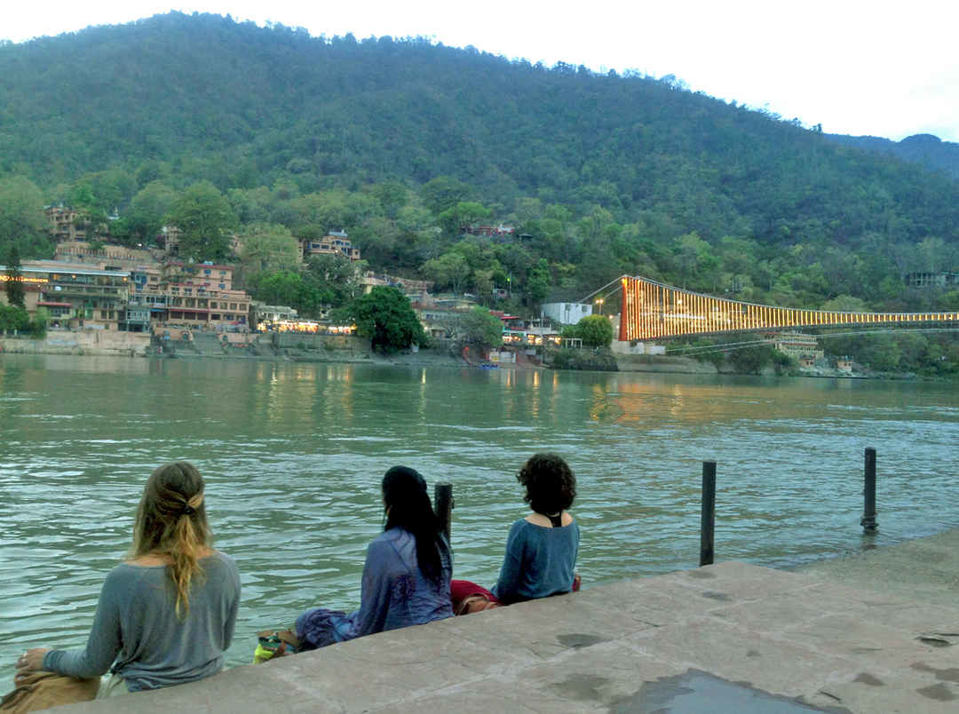 Three women sitting by the Ganges River in Rishikesh, with an illuminated suspension bridge and hill in the background, reflecting a peaceful solo travel experience