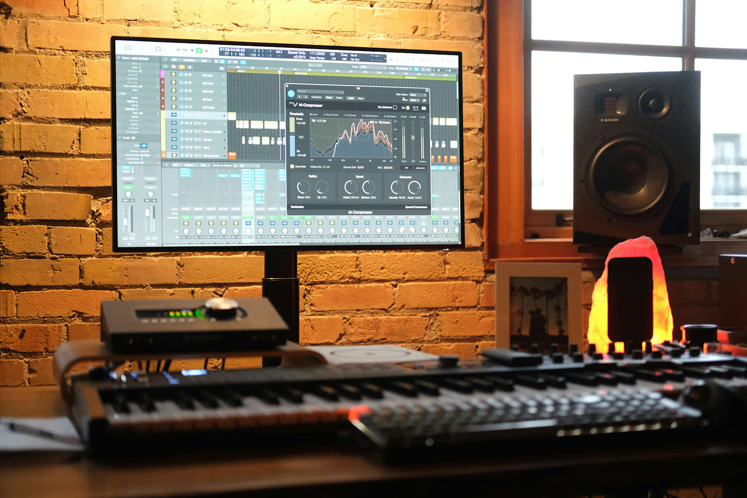A modern music production studio setup featuring audio mixing software on a monitor, MIDI keyboard, audio interface, and studio monitor speaker