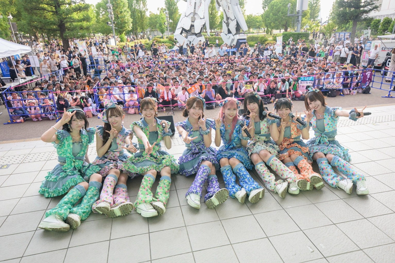Idol group posing with microphones during a live outdoor event in Odaiba with a large crowd of fans in the background.