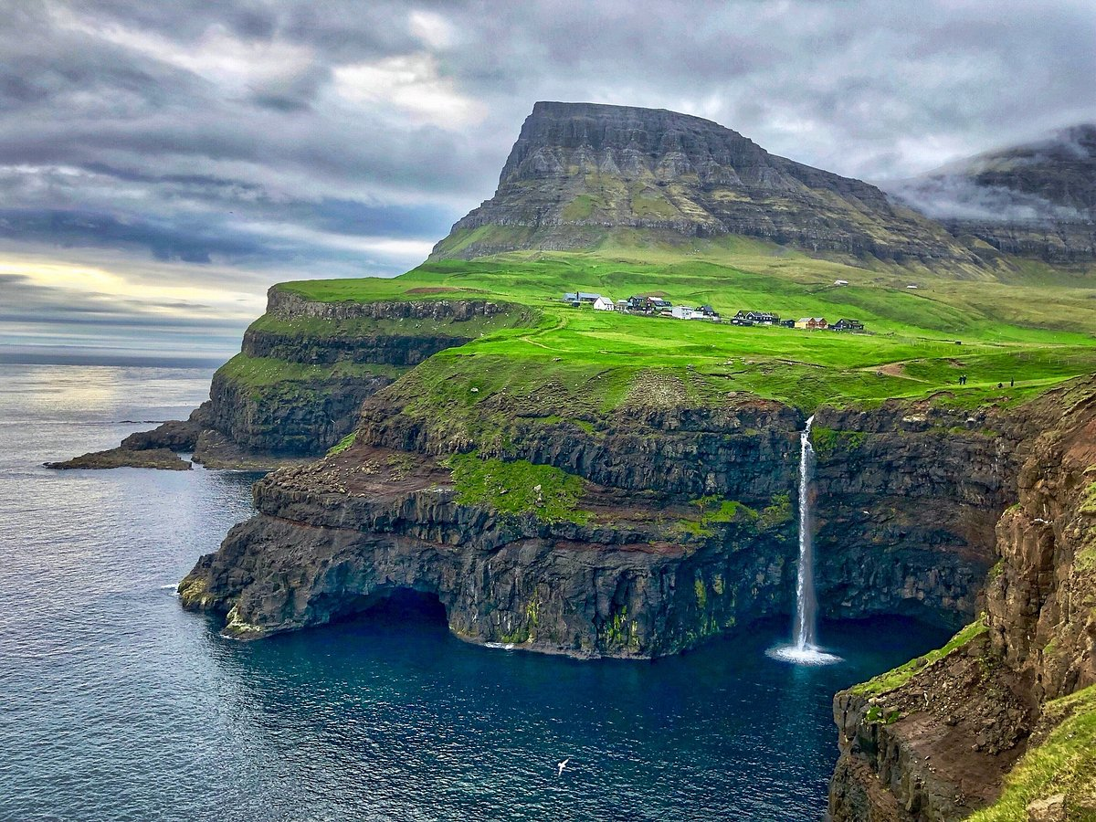 Mulafossur waterfall cascading into the ocean with lush green cliffs and mountain backdrop in the Faroe Islands