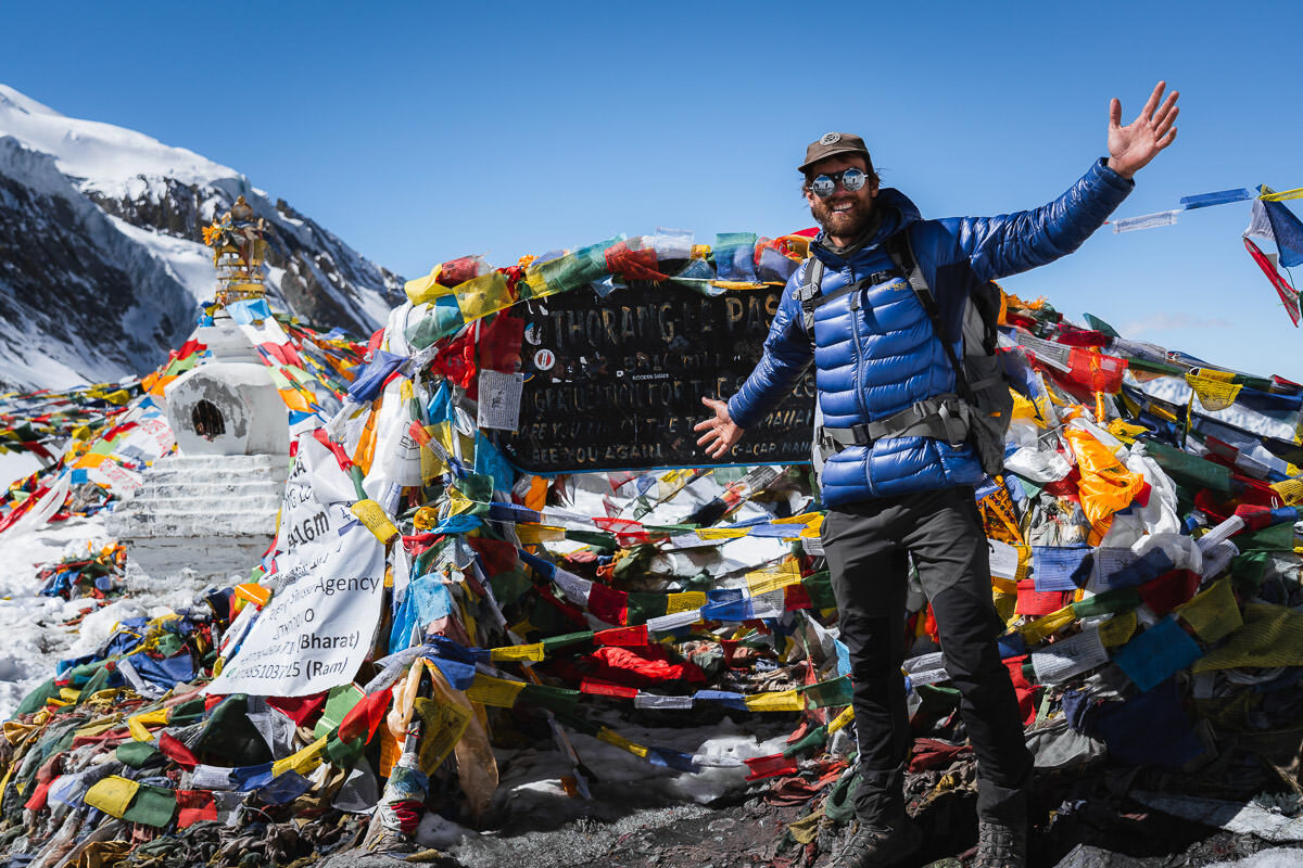 Trekker celebrating the accomplishment of crossing Thorong La Pass on the iconic Annapurna Circuit trek in Nepal, surrounded by colorful prayer flags and stunning Himalayan mountain views