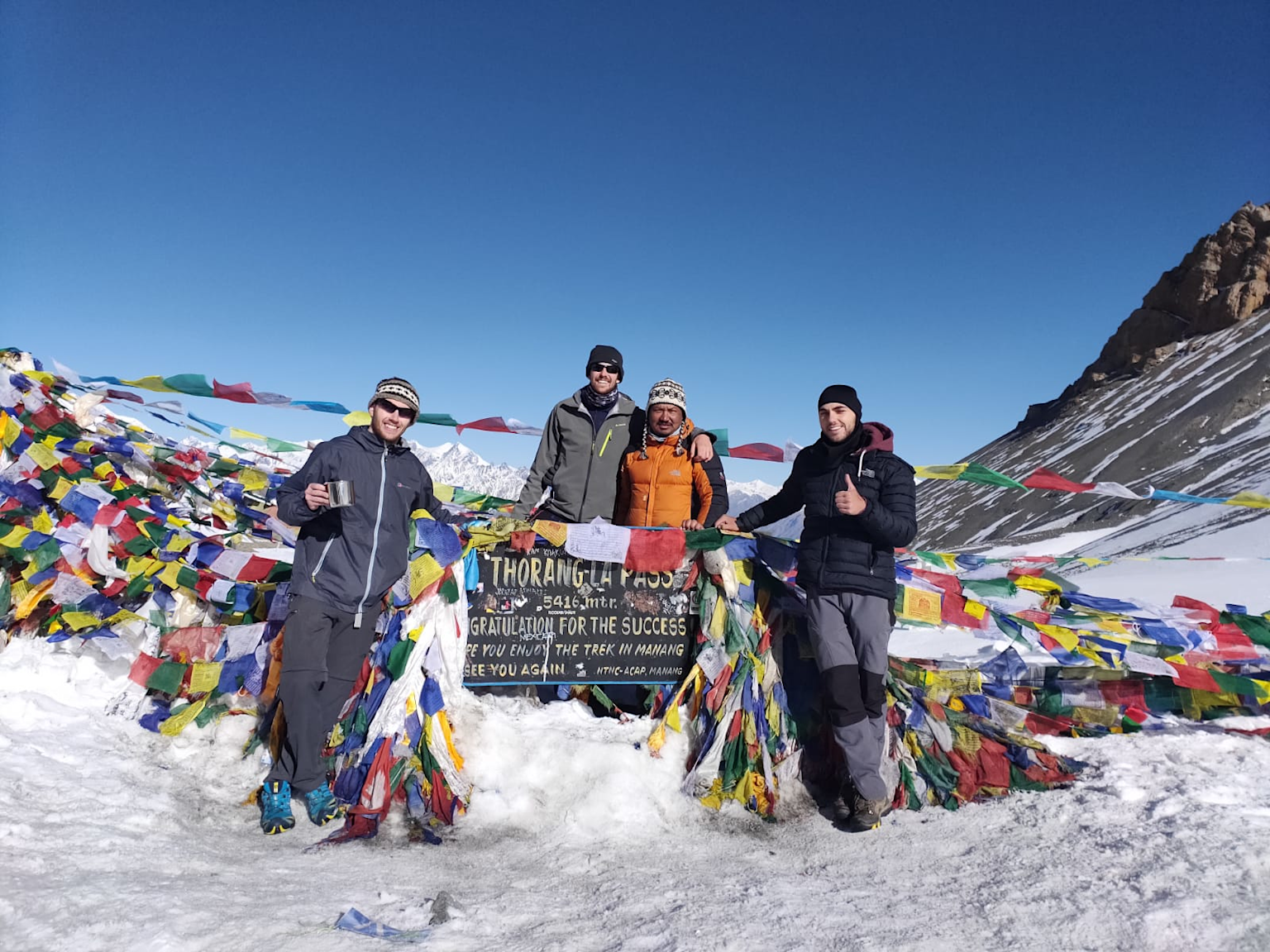 Trekkers celebrating their successful crossing of the Thorong La Pass at 5416 meters in Nepal, surrounded by traditional colorful prayer flags and stunning mountain scenery