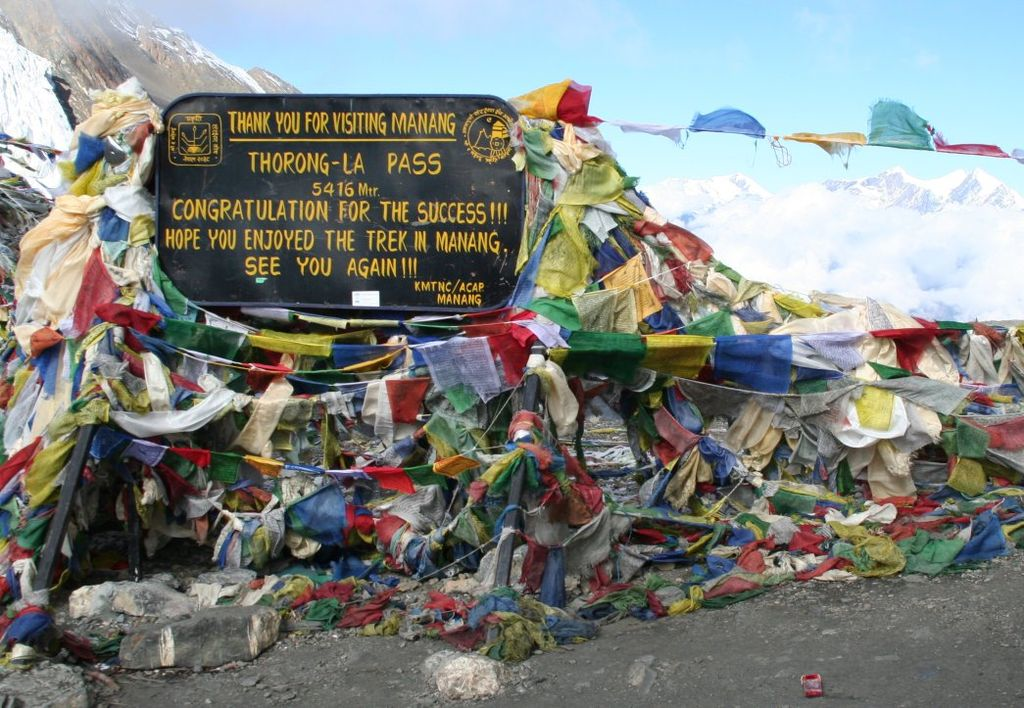 Signboard at Thorong La Pass (5416m), Nepal, surrounded by colorful prayer flags symbolizing trekking success and cultural heritage in the Himalayas