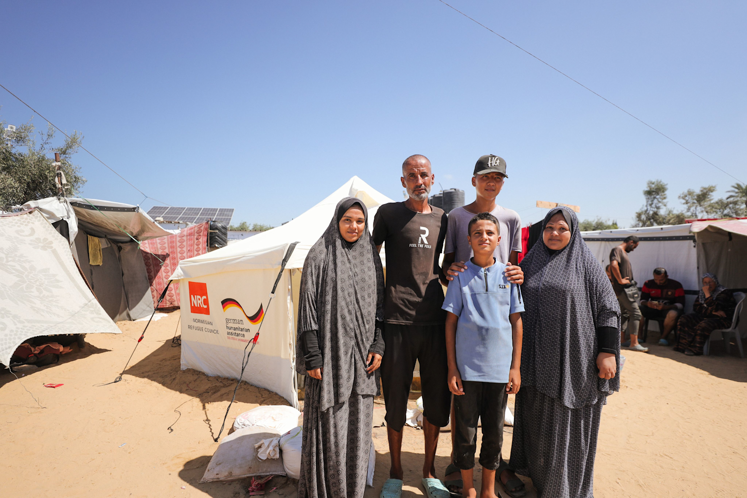 Displaced family posing in front of a humanitarian aid tent provided by the Norwegian Refugee Council and German assistance in a refugee camp