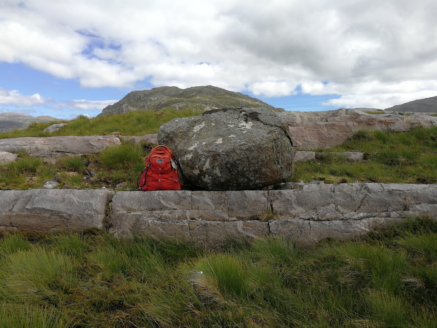 A large glacial erratic boulder resting on rocky terrain with a red backpack for scale, illustrating glacial erratic formations in natural landscapes