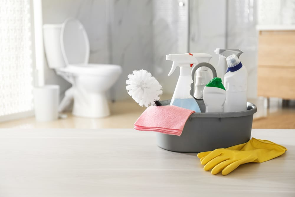 Bathroom cleaning supplies including spray bottles, a toilet brush, cleaning cloth, and gloves arranged in a modern bathroom setting
