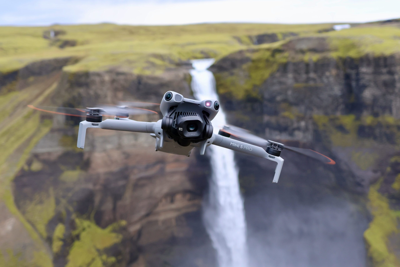 DJI Mini 5 Pro drone flying near a waterfall in a scenic landscape, illustrating its capability for high-quality aerial photography