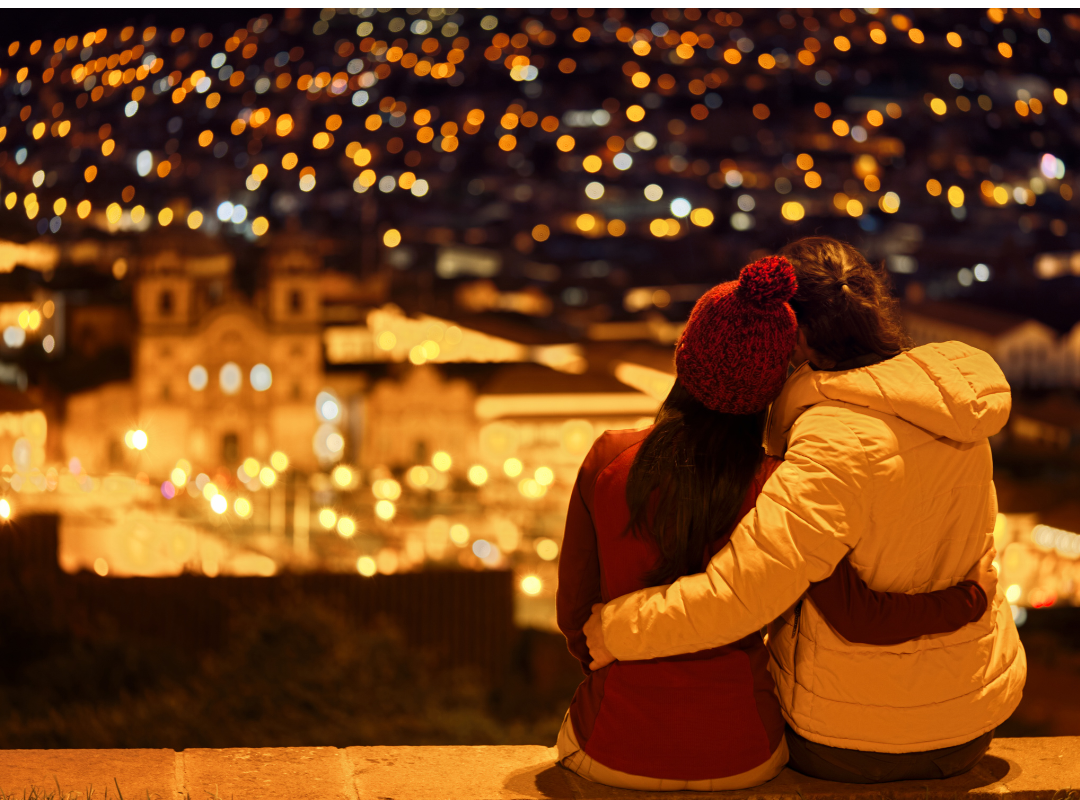 A couple embraces warmly as they enjoy a tranquil night view of a softly glowing cityscape, evoking a romantic and cultural travel experience likely in a historic city such as Cusco, Peru