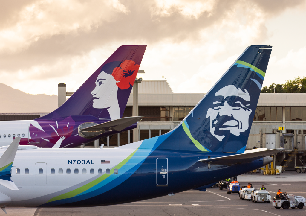 Hawaiian Airlines and Alaska Airlines planes parked side-by-side at an airport, showing the connection between the two carriers amid acquisition news