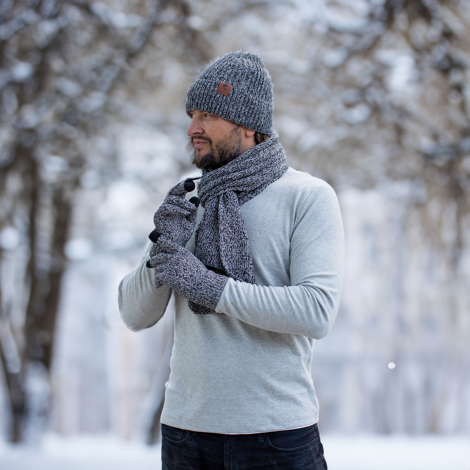Man wearing a coordinated set of gray knitted winter accessories including beanie, scarf, and gloves in a snowy outdoor setting.