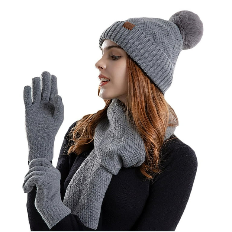 A woman wearing a coordinated set of grey knitted winter accessories including a beanie with pom-pom, scarf, and gloves.