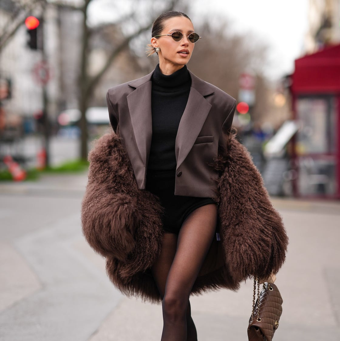 A woman wearing a chocolate brown oversized blazer and fluffy winter coat with black tights and a mocha quilted handbag, showcasing a 2025 winter fashion look for women.