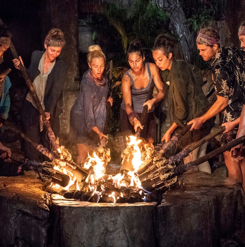 Survivor contestants participate in tribal council around a fire during a nighttime vote