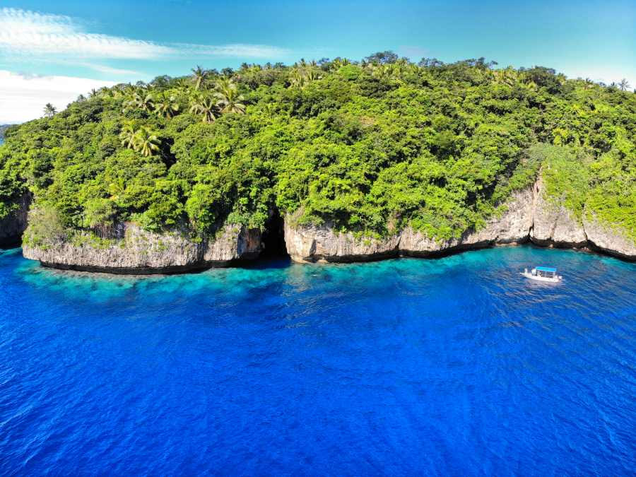 Aerial view of a green island with rocky cliffs and a boat in clear blue ocean waters, typical of a tropical diving location