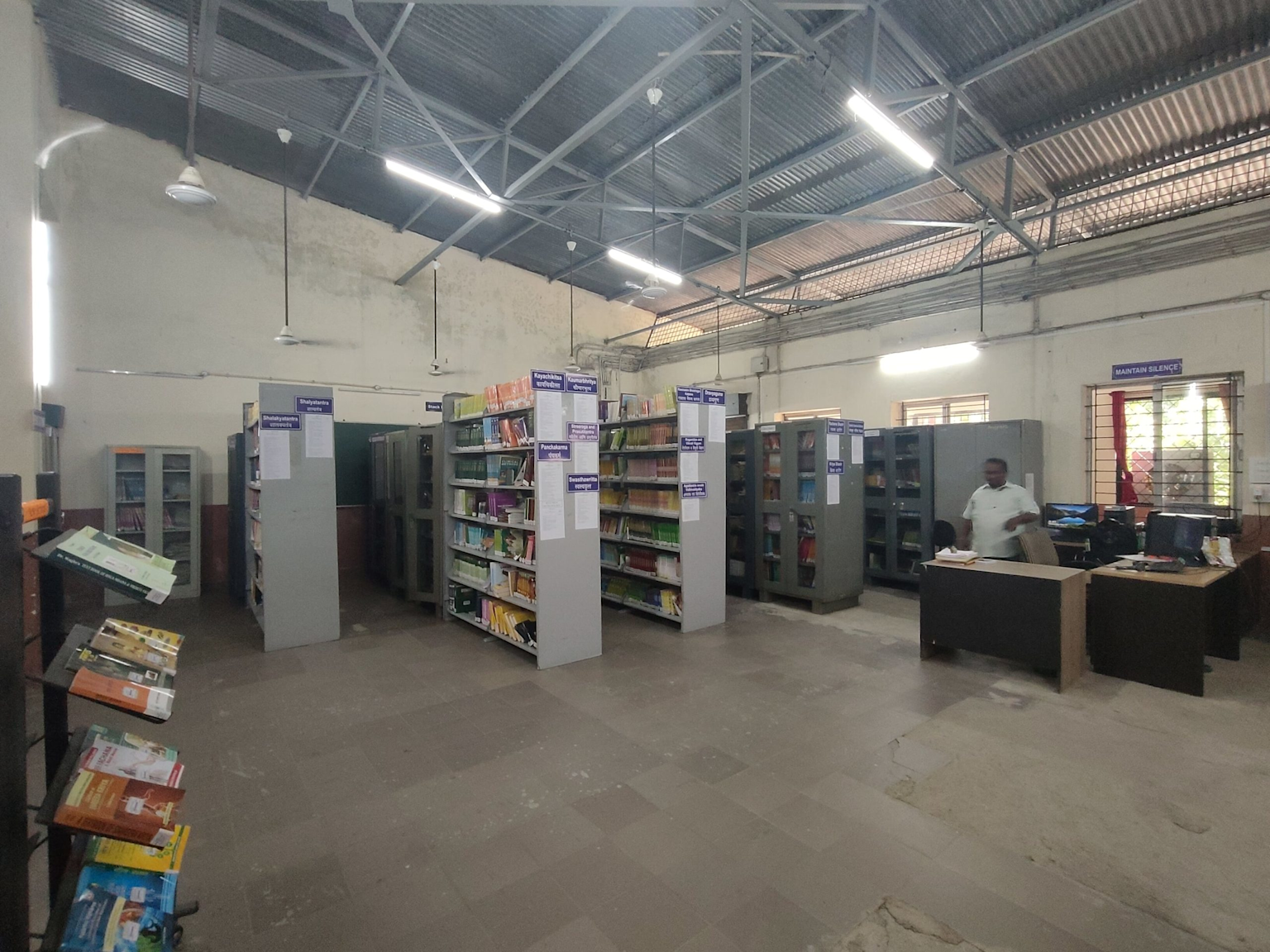 Interior of an Ayurvedic college library showing organized shelves by Ayurveda departments with books and a staff member